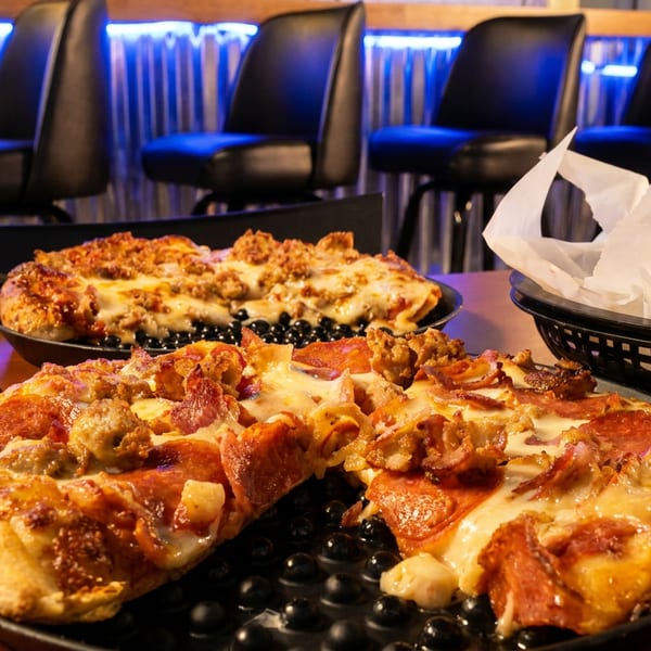 Two pizzas with various toppings sit on a table in a dimly lit restaurant with bar stools.
