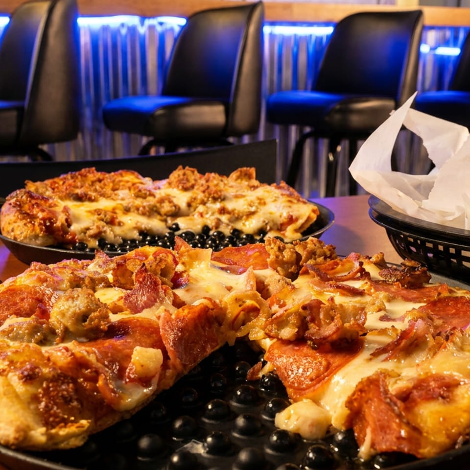Two pizzas with various toppings sit on a table in a dimly lit restaurant with bar stools.