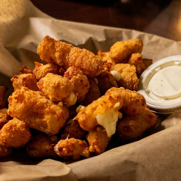 Golden-brown fried cheese curds with a side of dipping sauce in a paper-lined basket.